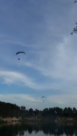 This vertical video captures a scenic view of a body of water with a dense forest lining its edge. Two paragliders are visible in the sky, soaring against a backdrop of a bright blue sky with scattered white clouds. The paragliders, one higher and closer to the foreground with a blue and white wing, and another further in the distance, suggest an activity of recreational flying or a competition. The sun appears to be shining, indicating a daytime setting, possibly late afternoon or early evening given the quality of light. The calm water reflects the trees and the sky, adding to the tranquility of the scene. The presence of a forest and water hints at a natural landscape, likely a lake or river. The overall mood is peaceful and adventurous, with the vastness of the sky contrasting with the grounded beauty of the forest and water. No visible text is present in the video. The location context of Sulejów, Poland, suggests this scene is likely taking place in a natural area around that region.
