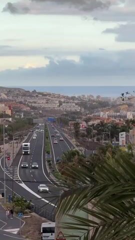 This is a vertical video capturing a view of a highway in Adeje, Spain. The highway stretches into the distance, lined with palm trees and leading towards a coastal town with buildings cascading down a hillside towards the ocean. The sky is overcast with dramatic clouds, suggesting either early morning or late evening, with the sun's light filtering through. 

The highway is moderately busy with cars and a large white truck. On the left side of the road, there's a pedestrian walkway with a few people walking, one of whom is carrying a bag. Further down the road, a white van is parked on the side. A prominent red and white sign is visible near the van, and further along the highway, there's a gas station with the "Disa" logo. The town in the background appears to be a tourist resort with multi-story buildings and the shimmer of the ocean under the diffused light. The foreground is dominated by the fronds of a large palm tree, partially obscuring the view. The overall mood is one of serene observation of daily life in a picturesque location.