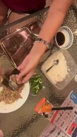 Here is a concise description of the video:

The video shows a person making temaki sushi.  They are adding rice and a tuna mixture to a sheet of nori seaweed. There are also cucumber slices in a bowl and soy sauce on the table. The scene is shot from directly overhead.  The person is wearing a red shirt and a beaded bracelet. A promotional flyer is on the table as well.