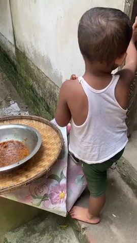 A barefoot toddler in a white tank top and green shorts stands on a step beside a table. The table is draped with a floral cloth, and on top is a woven tray holding a metal bowl filled with a brown substance. The child holds a small piece of the brown substance in their right hand, examining it.  Behind the child is a white wall. To the left is a narrow brick pathway bordered by a moss-covered wall.