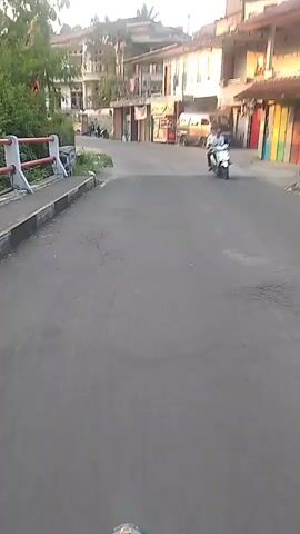 The video shows a first-person perspective of someone riding a motorcycle down a street in a village. The street is paved, and there are buildings on either side. The buildings appear to be residential homes or small businesses. There are trees and vegetation along the side of the road. In the distance, there are people riding motorcycles and a few parked vehicles. The sun is setting, casting a warm glow on the scene.
