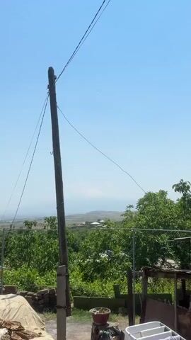 The video shows a clear blue sky with a tall wooden pole in the foreground. Wires are strung from the pole across the frame. In the background, there is a distant, snow-capped mountain range, partially obscured by haze. Below the mountains, a rural landscape with scattered houses and trees is visible. The foreground also shows some building materials, including a pile of sand and some bricks. A potted plant and laundry basket are also visible in the lower right corner. The overall impression is a sunny day in a rural setting with a prominent natural landmark.