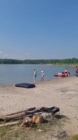 A sunny day at a lake beach. The water is calm and blue, with a treeline on the far shore. Several people are visible: a man in blue shorts walks into the water, followed by a child in a white hat. Further down the beach, a red jet ski is moored, and a group of people are gathered near it. An inflatable mattress lies on the sand near the water's edge. In the foreground, a fire pit with logs and a grill is set up on the sandy beach. The sky is clear blue with a few wispy clouds.