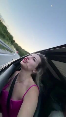A woman with blonde hair wearing a pink dress and a seatbelt sits in the passenger seat of a car with the sunroof open. She is yelling, smiling, and shaking her head. The car is driving on a winding road with a green hillside to the side. The sky is clear and blue.
