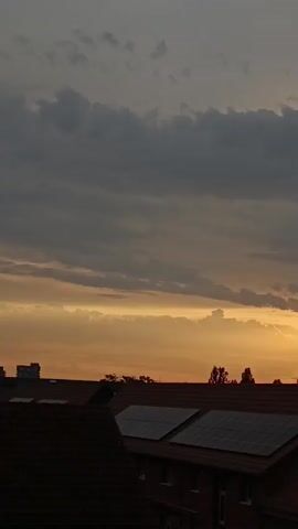 The video shows a sunset over a residential area. The sky is filled with dramatic clouds, with the sun peeking through them, casting a warm orange glow. In the foreground, the rooftops of buildings are visible, some equipped with solar panels. Silhouetted trees can be seen against the horizon. The overall impression is one of a serene and colorful end to the day.