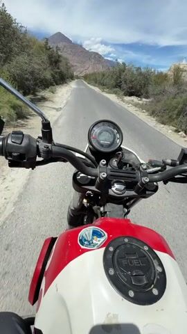 The video shows a first-person perspective of someone riding a motorcycle on a paved road. The rider is wearing a helmet and gloves. The road is surrounded by dry, rocky terrain with sparse vegetation and mountains in the background. The sky is blue with some clouds. The motorcycle is red and white. The rider briefly makes a peace sign to the camera. The video captures the motion of riding, with the scenery blurring slightly.