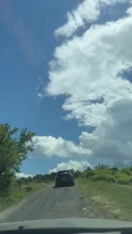 The video shows the view from inside a moving vehicle driving on a paved country road. A dark-colored car is driving ahead of the camera car. The road is surrounded by grassy fields with scattered trees and bushes. The sky is bright blue with fluffy white clouds. The windshield of the camera car is visible at the bottom of the frame.
