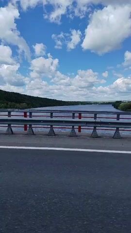 The video shows a view from a bridge looking out over a wide river under a blue sky with fluffy white clouds. The bridge has a guardrail made of metal poles with red horizontal bars. The road on the bridge is asphalt, with a white line marking the lane. Trees and hills line the banks of the river, and there are some buildings visible in the distance on the far bank. The water is calm and reflects the sky. The camera appears to be moving, suggesting the video was taken from a moving vehicle.