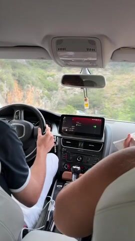 The video shows the interior of a car traveling on a road with a cliff face on the left and greenery on the right. A person's hands are visible on the steering wheel and gear shift, indicating they are driving. Another person is seated in the passenger seat, holding a white object in their hands. The car's infotainment system displays a navigation screen. The overall impression is of a journey through a scenic, mountainous area.
