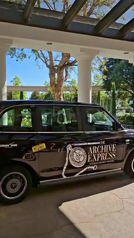The video shows a black London taxi parked under a covered patio with white columns and a glass roof. The taxi has "THE ARCHIV EXPRESS" written on its side, along with a logo of a chain and a compass. A large, colorful statue of a jester and another statue of a person in historical clothing are visible in the background, along with trees and a bright blue sky. In the foreground, a vintage beige convertible is parked. The scene appears to be outdoors, possibly at an amusement park or a historical site. There are no people clearly visible in the video.