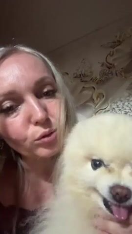 A woman with blonde hair holds a small, fluffy cream-colored dog in her arms. The woman is looking at the camera and speaking. The dog is looking at the camera with its mouth open and tongue sticking out, appearing to be smiling. The woman kisses the dog. The background is a wall with decorative wallpaper.
