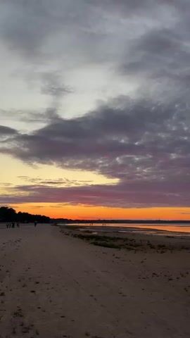 The video shows a beach at sunset. The sky is filled with colorful clouds, ranging from gray and pink to orange. The sun is setting over the water, casting a warm glow on the scene. There is a sandy beach in the foreground, with some vegetation growing near the water's edge. In the distance, a few people are walking along the beach. One person, wearing a pink shirt, is walking towards the water. The overall mood of the video is peaceful and serene.