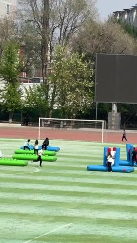 The video shows a group of people participating in an inflatable obstacle course on a green, striped field. The obstacles include green, filled cylinders, and structures with blue bases and colorful pillars. People are running, climbing, and navigating the obstacles, some falling in the process. In the background, there's a running track, a soccer goal, trees, and buildings. A large black screen stands tall behind the obstacles, and stone monuments with Chinese characters are visible next to it. The sky is clear and bright.