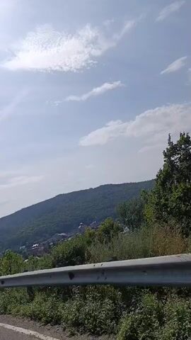 A first-person perspective video captures a drive through a rural landscape. The scene is dominated by lush green hills dotted with houses, suggesting a small village nestled within the natural scenery. A metallic guardrail runs along the bottom of the frame, indicating the presence of a road. The sky is a vibrant blue with scattered white clouds, suggesting fair weather. The video appears to be taken from a moving vehicle, evidenced by the slight blur and the changing perspective of the scenery. There are no people or animals visible in the frame. The video is short, lasting only a few seconds, and cycles through the same view.