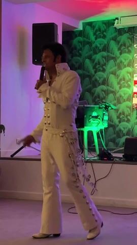 Here's a breakdown of the video:

A man dressed as Elvis Presley is singing into a microphone. He wears a white jumpsuit with large silver buttons and decorative embellishments, white shoes, and has a dark wig styled like Elvis. He’s standing on a low stage.

Behind him is a speaker on a stand, a backdrop with tropical foliage and star patterns, and a small table with equipment on it.

The setting is indoors, lit with pink and purple lighting. Other people are milling about the room in the background, some seated in chairs, and houseplants are visible. He gestures and smiles while singing.