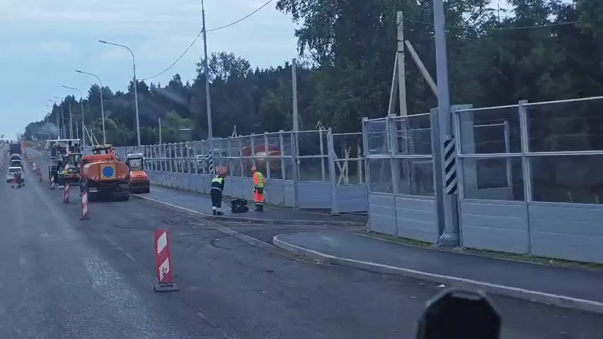 The video captures a construction site on a road, likely for resurfacing or repair. Several pieces of heavy machinery are present, including an orange water truck, a paver, and at least two bulldozers or compactors. Two construction workers, dressed in high-visibility safety vests and orange work pants, are seen operating a piece of equipment on the paved surface. They appear to be compacting or smoothing the road material. Traffic cones are placed along the road to direct traffic and cordon off the work area. A transparent noise barrier is erected on the right side of the road, separating the construction zone from a wooded area. The sky is overcast, suggesting a cloudy day, and the overall lighting indicates it's either early morning or late evening. The scene is dynamic, with ongoing construction work.