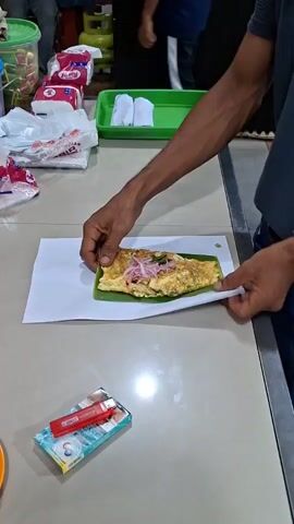 A person's hands are shown wrapping a food item in white paper. The food item appears to be on a green leaf and has a golden-brown cooked exterior. The person is using their right hand to fold the paper around the food and their left hand to steady it. A watch is visible on the left wrist. In the background, there are several packs of tissues, a green plastic tray with white folded items, and a pack of cigarettes with a red lighter on a table. The environment appears to be indoors, possibly a food stall or market.