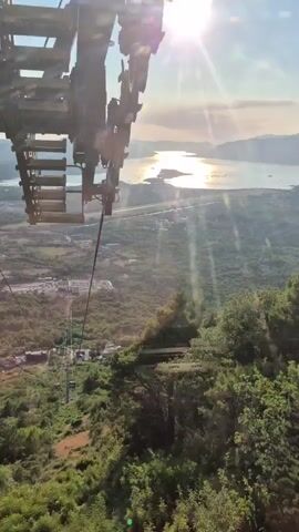 The video is a first-person perspective from a cable car ascending a mountain. In the foreground, the top of a tall support tower for the cable car system is visible, with the cable stretching upwards towards the sun, which is high in the sky and creating lens flares. The cable car itself is moving away from the viewer, with the side of one car briefly visible. Below and to the left of the tower, more cable cars can be seen moving along the wire further down the mountain. The landscape is lush green, covered in trees and vegetation, with the terrain sloping downwards. In the distance, a body of water, likely a bay or sea, reflects the sunlight. A road or highway runs along the coastline, and there are some scattered buildings and structures visible on the land between the mountain and the water. The sky is clear blue with a few wispy clouds.