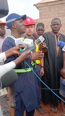 A group of men are being interviewed by the press outdoors. The speaker is a man with a navy cap and a reflector vest, surrounded by several microphones from news organizations. Behind him are other men, including one with a red hard hat, one in traditional Islamic garb, and one holding an “ARISE News” microphone. To the left of the speaker, other men watch. A small child can be seen in the lower right. The background includes a brick wall and what seems to be a rooftop. Overall, the scene appears to be a press event after or during some kind of site visit.