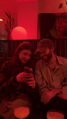 A young woman and a young man are sitting next to each other at a table in a dimly lit room with red lighting. The woman is looking at her phone, while the man looks at the camera and smiles. On the table in front of them are two cocktails. Behind the man is a black speaker and some plants. There is a light fixture in the shape of a ball behind the woman.