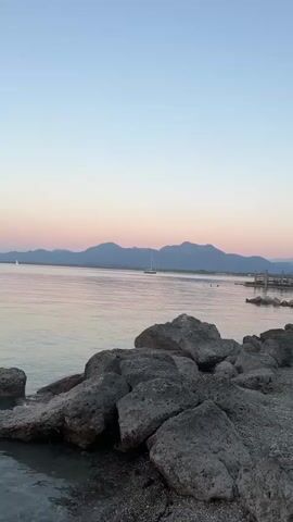 A video shows a peaceful scene at dusk. The sky transitions from a pale blue to a soft pink and orange hue. The water of a lake or sea is calm and reflects the colors of the sky. In the foreground, a rocky shoreline is visible, with several large, grey rocks. Further out on the water, a sailboat with its mast is sailing. In the mid-ground, several people are swimming or wading in the water. To the right, a wooden pier or dock is partially visible. The overall atmosphere is serene and tranquil.