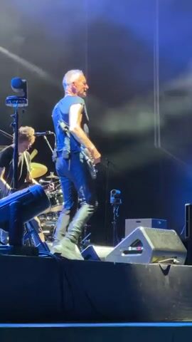 The video shows a man with short, grey hair playing a guitar on a dimly lit stage. He is wearing a dark blue t-shirt and dark pants. The stage is equipped with musical instruments, including drums in the background, and speakers. Blue stage lighting illuminates the scene. The man is facing away from the camera, looking to his right, and appears to be in the middle of a performance.