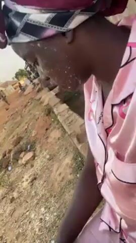 The video shows a woman in a pink pajama set and a head wrap standing in a dusty, rural area. Her face appears to have a white substance on it. The camera pans around to show a dirt road with houses made of mud bricks and corrugated iron roofs. People are seen walking and standing around in the background. The scene is set in an impoverished area.
