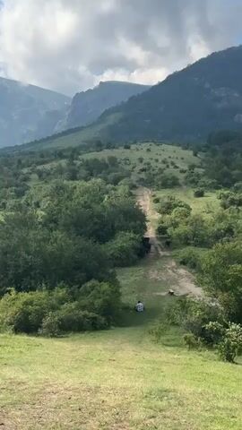This video captures a picturesque mountainous landscape with lush green vegetation under a partly cloudy sky. The scene appears to be set in Kalofer, Bulgaria.

In the foreground, a grassy slope leads down towards a dirt path winding through dense trees and bushes. A person in a white and black checkered shirt is seated on the grass near the path, observing the surroundings. Further along the path, a small herd of dark-colored animals, possibly horses or cattle, are moving.

The middle ground is dominated by rolling hills covered in a rich tapestry of green foliage, with a mix of trees and shrubs. A discernible dirt track cuts through this verdant expanse, leading towards the background.

The background features majestic, imposing mountains, their peaks partially shrouded in mist or clouds. The largest mountain in the distance has a prominent tower or structure on its summit. The sky is a mix of bright blue and dramatic, billowing white and grey clouds, suggesting a dynamic weather pattern.

The time of day appears to be late morning or early afternoon, given the bright sunlight illuminating the landscape. The overall atmosphere is one of tranquility and natural beauty, with a sense of peaceful coexistence between humans, animals, and the environment. No visible text is present in the video.