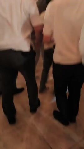 This video depicts a lively celebration where a man is being lifted and carried on the shoulders of other men. Several individuals are wearing white shirts and kippahs, suggesting a Jewish cultural or religious gathering. In the background, there is a stage with lights and a large screen displaying illuminated patterns. The atmosphere appears festive, with people actively participating in the celebratory activity. The camera focuses on the man being carried, who is wearing a suit jacket and a kippah. A young boy is also visible on someone's shoulders, near the man being carried. The lighting suggests it is evening or indoors with artificial lighting.