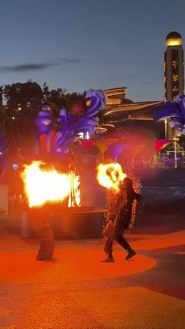 The video shows a fire performance outdoors at dusk. Two individuals are on a paved circular area, each spraying large plumes of fire from their hands. The fire is bright orange and yellow and creates a lot of smoke and sparks. In the background, there are some buildings with lights, and trees. The sky is a dark blue with some clouds. Several people are watching the performance, and some are holding up their phones to record it. The ground is marked with colorful patterns, including stars.