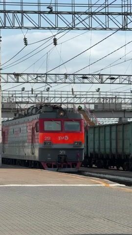 A red and gray passenger train is parked at a train station. The train is being lit by the sun. The train is next to the platform, which is made of stone. There is a bridge above the train track.