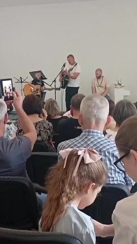 A video clip shows an indoor performance with an audience seated and three men on a stage. The man in the center is standing and playing an acoustic guitar while singing into a microphone. To his left, a younger man is also standing and playing an acoustic guitar. To the right of the singer, another man is seated. The stage is minimally decorated with some plants. The audience is facing the stage, with some individuals recording the performance on their phones. The room appears to be a white-walled hall.