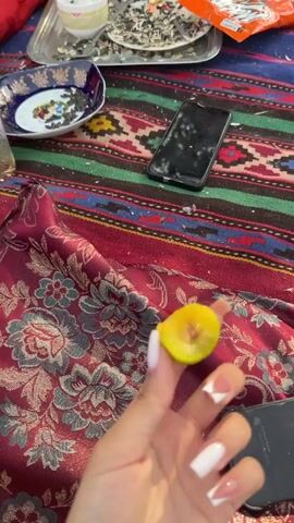The video opens with a close-up of a hand with white French manicured nails holding a cut piece of yellow fruit. A cell phone lies on a richly patterned red and blue textile in the background. The scene shifts to a woman sitting on the same textile, looking surprised. In front of her are various items including a large metal bowl, glasses, a mug, a plastic bag, and a bag of snacks. A second cell phone is visible in the foreground. The next shot shows a woman in a green t-shirt and blue pants with a patterned sash around her waist, looking upwards with a pained expression. She has a braid of dark hair. The final segment shows a young person in a black t-shirt and light-colored shorts looking at the camera, with the legs of someone wearing red pajama pants with a black and red lip pattern in the foreground. Behind them, a duck is visible. The setting appears to be outdoors, with trees, vegetation, and hilly terrain in the background, suggesting a rural or natural environment.