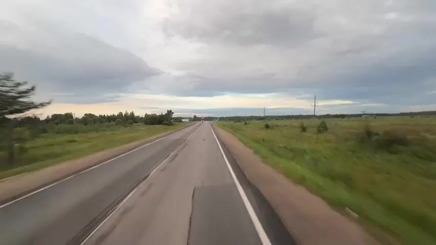 The video shows a forward view from a vehicle traveling on a rural highway. The road is paved with asphalt and has white lane markings. On either side of the road are grassy fields with scattered trees and bushes. In the distance, there are more trees and some utility poles. The sky is cloudy with hints of sunset colors. A red truck with a white trailer is seen approaching and passing the vehicle from the opposite direction. A few signs are visible along the roadside, including a diamond-shaped sign with a red border and a white triangle inside, and a blue rectangular sign with white arrows. The overall environment suggests a somewhat overcast day in a rural setting.