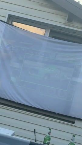 The video shows an outdoor scene with a swimming pool in the foreground. A house is visible in the background, with a large white screen hung on its side. The screen displays the words "Lightning McQueen" in large letters. Two bottles of beer are placed on a table near the pool. A person is partially submerged in the pool, facing away from the camera. The sky is clear and blue. The camera pans slowly across the scene, capturing the view of the house, screen, pool, and person.