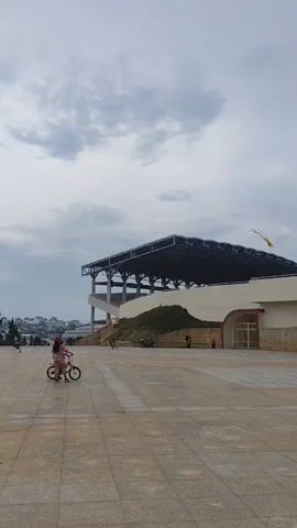 Here is a description of the video:

The video shows a large, paved square in front of what appears to be a stadium or covered outdoor arena. The structure has a large metal roof and a concrete base. There's a grassy area built into the base of the structure, and a large electronic display screen is visible on the right side. A young child wearing a helmet and riding a small bicycle is in the foreground. Other people are visible in the distance near the edge of the square. The sky is overcast.