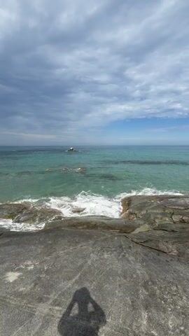 A vertical, wide-angle shot of the ocean from a rocky shore. The water is a vibrant turquoise and the sky is filled with a mix of blue patches and dark gray clouds. Waves crash against the rocks in the foreground and on a small island in the distance. The shadow of a person holding a phone up to take the picture is visible in the bottom center of the frame. The rocks in the foreground appear to be dark gray and textured, with some sand visible.