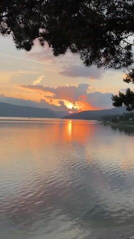 A vertical time-lapse video shows a sunset over a body of water. The sun, a bright orange circle, descends behind a distant mountain range, casting a golden reflection on the water. The sky transitions from light orange to a darker, more intense orange and red as the sun sets. Dark clouds move across the sky, partially obscuring the sun at times. Silhouetted trees are visible in the foreground, framing the shot from the top and sides.
