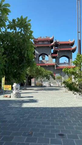 The video pans across an outdoor courtyard in front of a temple. The courtyard is paved with grey square tiles. In the foreground, there is a large green tree with lush leaves. To the left, a woman wearing a white hat and a blue shirt is sitting on wooden steps, looking at her phone. Behind her, there is a white statue of an animal. Further back, there is a large, ornate temple structure with a grey stone facade, red tiled roofs, and large archways. A tall, white statue of a person, likely a Buddha, stands prominently on top of the temple roof, silhouetted against a clear blue sky. There are other trees and foliage around the temple. A yellow sign with Vietnamese writing is visible near the woman.