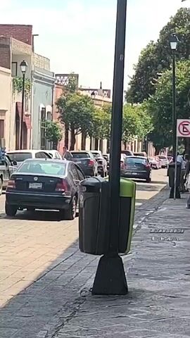 The video shows a street scene in what appears to be a historic district, with cobblestone paving and buildings featuring traditional architecture. Various cars are parked along the side of the street. A light-colored SUV is parked near a restaurant called "SWEXICAN TAQUERIA & BURGERS". Several people are standing and talking on the sidewalk near the restaurant. There is a trash bin attached to a lamppost in the foreground of the initial shot. The video pans slightly to the left, focusing on the group of people and the restaurant. The weather appears to be sunny and clear.