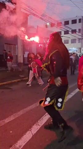 This is a shaky, vertically oriented video filmed at street level during what appears to be a parade or procession at dusk or dawn. The foreground is dominated by a person in a striking costume. This individual wears a black outfit with a red sash and decorative trim, black pants, and dark shoes. Their most prominent feature is a long, dark, possibly wig-like headdress that covers their head and shoulders, obscuring their face. They are holding a bright red flare in their right hand, which emits a significant amount of smoke and light.

To the left and behind this main figure, other participants are visible. Some are wearing masks and colorful, elaborate costumes, like a person in a vibrant pink outfit. Others are dressed more casually in everyday clothing and are observing the event. Several individuals in the middle ground are holding musical instruments, including what look like drums, and appear to be part of the procession.

The background shows a street lined with buildings, some of which have multiple stories and windows. Streetlights are visible, indicating it is not full daylight. The overall atmosphere is festive and dynamic, with the red flare casting a strong, dramatic glow on the scene. The ground has speed bumps and painted white lines, typical of a street. The camera movement is unsteady, suggesting it's being held by someone walking or moving with the procession.