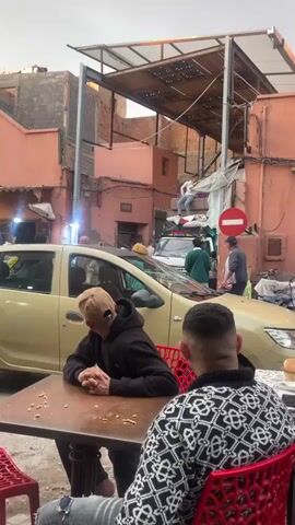 The video shows a street scene in what appears to be a densely populated urban area with narrow streets. The buildings are mostly two to three stories tall and painted in shades of pink and beige.  Overhead, a corrugated metal roof covers part of the street, offering partial shade. In the foreground, two men are seated at a table outside what appears to be a cafe. One man wears a brown baseball cap and a black hoodie, his head lowered. The other man wears a black and white patterned sweater and sits facing away from the camera. Behind them, a yellow taxi is parked, and beyond that, there is a white van loaded with what appears to be mattresses and luggage.  A man in a white shirt and blue jeans is sitting on top of the load, seeming to adjust the items. Several other people are standing near the van, some conversing with each other. A red "do not enter" sign is visible on a pole next to the van. The scene is busy with people and activity.