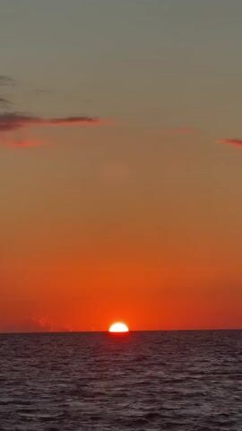 A video of a sunset over the ocean. The sun is a bright orange orb half-submerged below the horizon, casting a warm glow across the sky. The sky is a gradient of orange and pink, with a few wispy clouds. The ocean is a deep blue, with gentle waves. The video is static, with no camera movement.