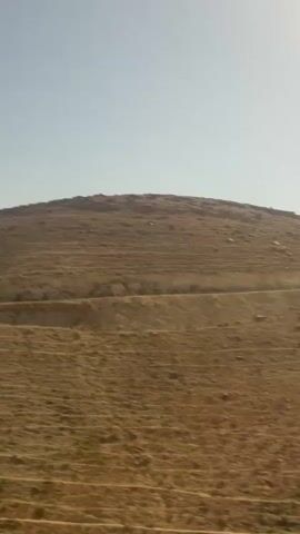 The video shows a panoramic view of a desert landscape with rolling hills and a clear sky. The terrain is dry and arid, with sparse vegetation, suggesting a hot and sunny climate. The time of day appears to be late morning or early afternoon, as indicated by the bright sunlight casting shadows on the hills. The location is Arad, Israel, a city known for its desert environment. There are no people or specific objects visible in the footage, focusing solely on the natural scenery. The overall mood is one of vastness and tranquility, characteristic of desert landscapes. No text is visible in the media file.