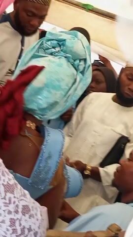 Here is a concise description of the video:

The video shows a group of people gathered closely together, possibly at an event or celebration. A woman with red hair and wearing a light blue dress with beaded trim is seated among them. There's a person in white lace next to her. A white sheet is held up, obscuring the view of some of the individuals. Other people are looking on and interacting with those in the immediate group. The scene is brightly lit.