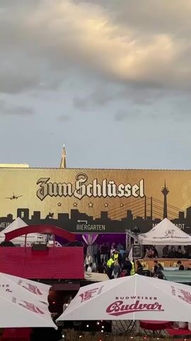 The video shows a bustling outdoor event. Above, a large gold sign reads "Zum Schlüssel BIERGARTEN" in ornate lettering, with a black silhouette of a city skyline below it. People are gathered in front of an entrance draped with purple and silver fabric. Some individuals are wearing high-visibility vests, possibly security personnel. In the foreground, white umbrellas with red lettering, one clearly showing "Budweiser," are set up. The overall atmosphere appears to be lively and crowded, suggesting a public gathering or festival. The sky is overcast with hints of light.