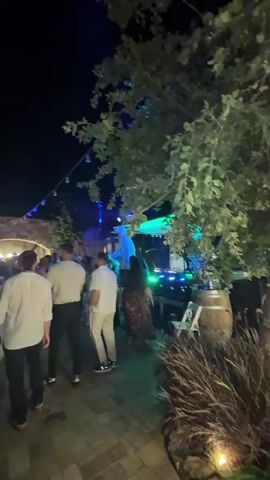 This is a video clip featuring a nighttime outdoor event, likely a celebration or party. The scene is illuminated by colorful artificial lights, including blue and green hues, casting a vibrant atmosphere. In the foreground, several individuals are visible, some facing away from the camera. One person in the foreground is wearing a white collared shirt. The background shows a gathering of people, suggesting a lively event. String lights are visible overhead, adding to the festive ambiance.

The event appears to be taking place in an outdoor space with paved ground and some foliage or greenery. There's a hint of architectural structures, possibly an arched entrance or a building facade, in the background. The overall mood seems to be one of enjoyment and social interaction, with people mingling and possibly dancing. The time of day is clearly night, as indicated by the dark sky and the artificial lighting. While the specific nature of the event isn't explicitly stated, the combination of lights, music (implied by the party atmosphere), and people suggests a festive occasion. No visible text is present in the video clip.
