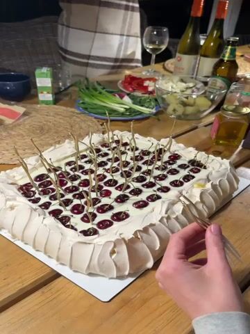 The scene is a wooden table set for a social gathering, illuminated by indoor lighting. A woman in a light grey sweatshirt is the main subject, seen meticulously preparing small, decorative gold-colored forks. She is separating and arranging them, her hands busy with the task. On the table, a large square cake with a cream topping, arranged cherries, and the same type of decorative forks stuck in the top is the centerpiece. Surrounding the cake are various food and beverage items: bottles of wine and beer, a jar of honey, and bowls filled with salad, shredded carrots, and other snacks. A glimpse of additional people can be seen around the table, one partially draped in a patterned blanket. The atmosphere suggests a relaxed and informal event, likely a celebration or get-together.