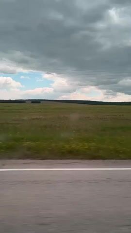 The video shows a view from a moving vehicle. The foreground consists of a road surface with a white line. Beyond the road is a vast, green field with scattered yellow wildflowers. In the distance, a dark line of trees marks the horizon. The sky is dramatic, with a mix of dark, heavy clouds and patches of bright blue sky with white clouds. There are no people or animals visible. The overall impression is a rural landscape under a dynamic sky.