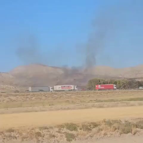 The video shows a view of a highway with several trucks driving on it. In the background, smoke is rising from hills, indicating a fire. One truck in the foreground has its door open, and a person wearing a brown jacket is visible in the driver's seat, appearing to be looking towards the smoke. The environment appears to be dry and rural, with hills and fields. The sky is clear and blue.